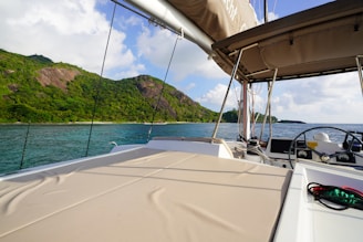 A sailboat cockpit with a cushioned seating area overlooks a beautiful seascape. Lush green hills rise in the background under a partly cloudy blue sky. The boat's canopy provides shade, and the steering wheel is visible on the right.