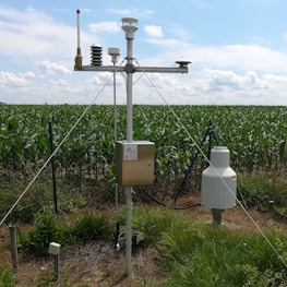A meteorological station is set up in a field of green crops, likely corn, under a partly cloudy sky. The station has several sensors and instruments mounted on a tall pole, secured with metal wires. Grassy vegetation surrounds the station, and a wire fence is visible in the background.