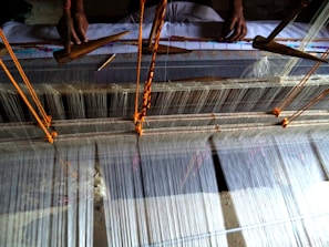 A traditional loom in use for weaving fabric. The loom is set up with numerous strands of thread, with two hands visible operating wooden shuttles to weave the threads together. The setup includes orange strings to manage the tension and alignment of the weaving threads.