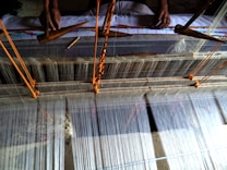 A traditional loom in use for weaving fabric. The loom is set up with numerous strands of thread, with two hands visible operating wooden shuttles to weave the threads together. The setup includes orange strings to manage the tension and alignment of the weaving threads.
