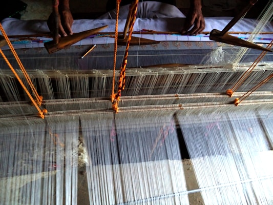 A traditional loom in use for weaving fabric. The loom is set up with numerous strands of thread, with two hands visible operating wooden shuttles to weave the threads together. The setup includes orange strings to manage the tension and alignment of the weaving threads.