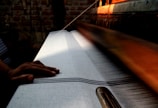 A hand rests on a weaving machine, where a white fabric with fine stripes is being produced. The setting is dimly lit, with warm light highlighting the machinery and creating a focus on the weaving process. The background features a brick wall.