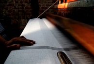 A hand rests on a weaving machine, where a white fabric with fine stripes is being produced. The setting is dimly lit, with warm light highlighting the machinery and creating a focus on the weaving process. The background features a brick wall.