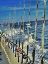 A line of fishing rods is neatly arranged along the railing of a boat, with the ocean and frothy waves visible in the background under a clear blue sky. A few pieces of clothing or towels are draped over the railing.
