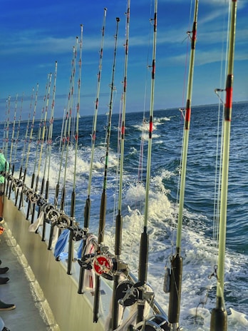 A line of fishing rods is neatly arranged along the railing of a boat, with the ocean and frothy waves visible in the background under a clear blue sky. A few pieces of clothing or towels are draped over the railing.