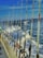 A line of fishing rods is neatly arranged along the railing of a boat, with the ocean and frothy waves visible in the background under a clear blue sky. A few pieces of clothing or towels are draped over the railing.