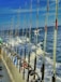 A line of fishing rods is neatly arranged along the railing of a boat, with the ocean and frothy waves visible in the background under a clear blue sky. A few pieces of clothing or towels are draped over the railing.