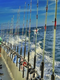A line of fishing rods is neatly arranged along the railing of a boat, with the ocean and frothy waves visible in the background under a clear blue sky. A few pieces of clothing or towels are draped over the railing.