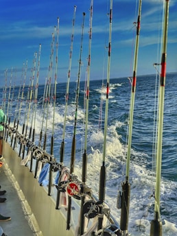 A line of fishing rods is neatly arranged along the railing of a boat, with the ocean and frothy waves visible in the background under a clear blue sky. A few pieces of clothing or towels are draped over the railing.