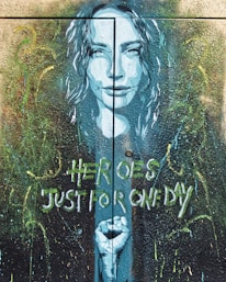 A mural artwork depicts a woman's face in a blue hue with her hair flowing around her. Below her face, a clenched fist is portrayed. The background is filled with abstract splatters and brushstrokes in various colors. The words 'Heroes Just for One Day' are painted in a stylized font.