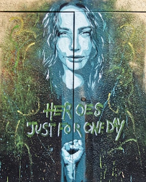 A mural artwork depicts a woman's face in a blue hue with her hair flowing around her. Below her face, a clenched fist is portrayed. The background is filled with abstract splatters and brushstrokes in various colors. The words 'Heroes Just for One Day' are painted in a stylized font.