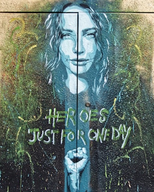 A mural artwork depicts a woman's face in a blue hue with her hair flowing around her. Below her face, a clenched fist is portrayed. The background is filled with abstract splatters and brushstrokes in various colors. The words 'Heroes Just for One Day' are painted in a stylized font.
