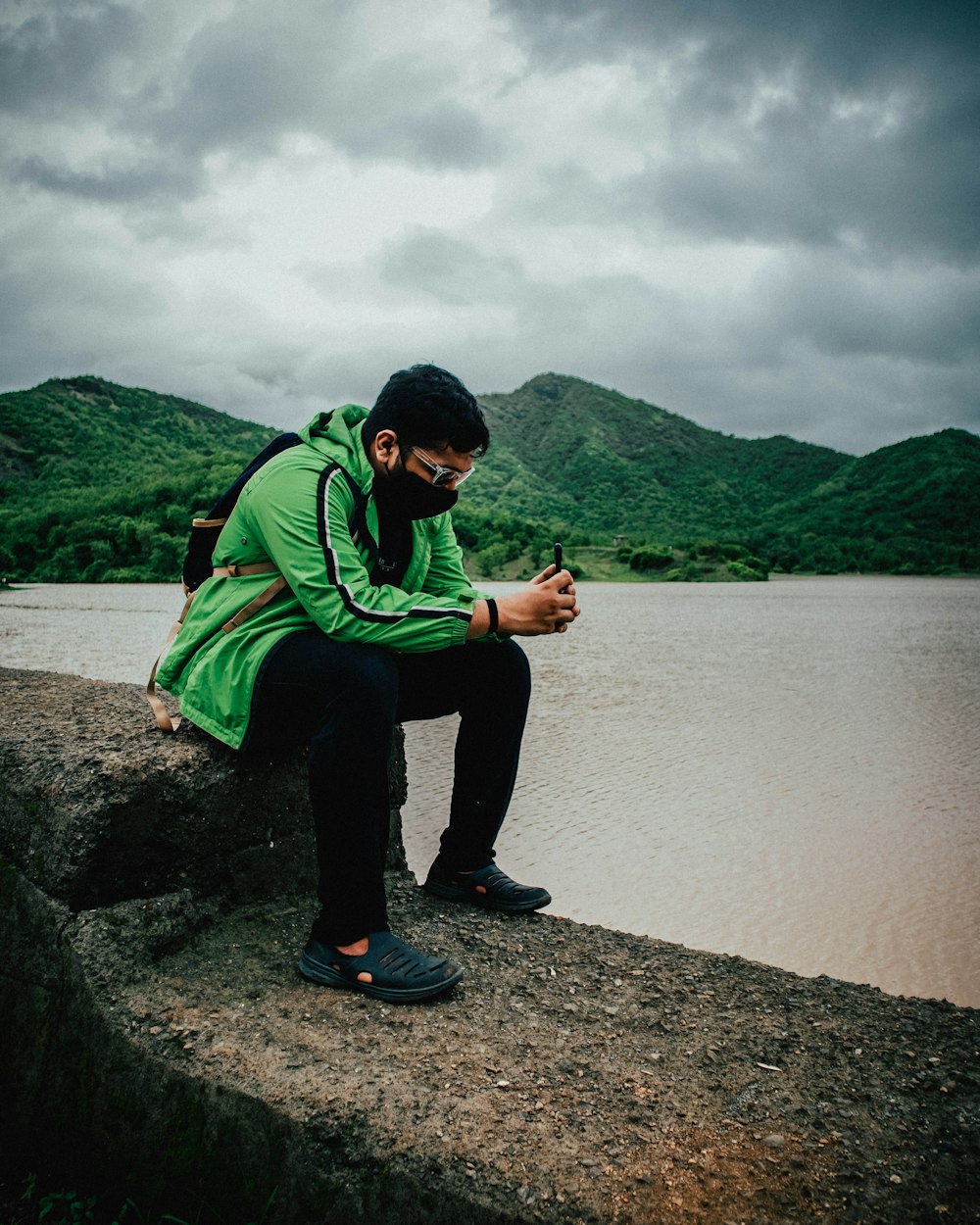 man in green hoodie and black pants sitting on gray rock near body of water during