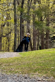 A person is cleaning up litter in a wooded area, using a large black trash bag. They are wearing a blue shirt and have a black jacket with yellow accents. The background features numerous tall trees with fresh green leaves, indicating it is spring.