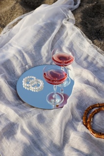 Two glasses of red wine placed on a round mirror, surrounded by a white sheer fabric. A string of pearls is elegantly positioned beside the glasses, and a wooden bracelet is visible nearby. The setup has an artistic and elegant flair, suggesting a leisurely, upscale picnic or decorative arrangement.