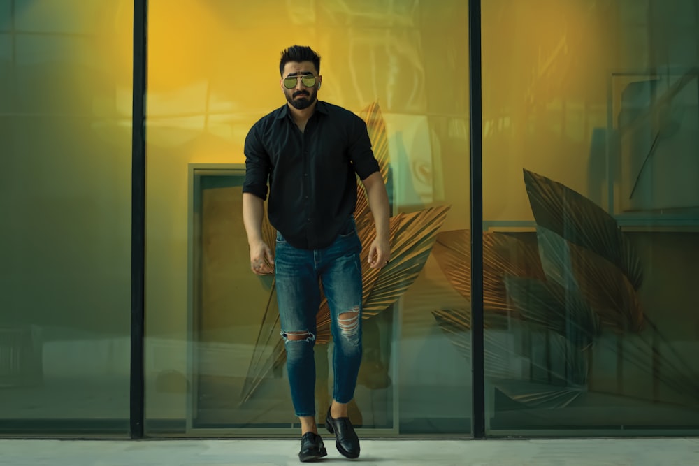 man in black polo shirt and blue denim jeans standing beside glass wall