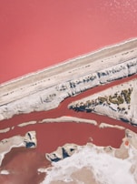 Aerial view of a vibrant, pink-colored body of water bordered by lighter sandy or rocky terrain. There are distinct paths or divisions of land cutting through the water, and variations in color suggest different depths or salinity levels. Sparse vegetation is visible in some areas.