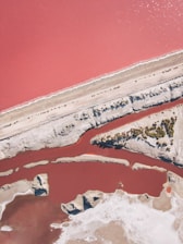 Aerial view of a vibrant, pink-colored body of water bordered by lighter sandy or rocky terrain. There are distinct paths or divisions of land cutting through the water, and variations in color suggest different depths or salinity levels. Sparse vegetation is visible in some areas.