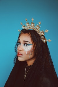 A person is adorned with a gold, ornate crown featuring intricate details and pearls. Their makeup includes striking silver tears and bold eye makeup, creating an artistic and dramatic appearance. A black lace veil partially covers their braided hair, and the background is a solid, calming blue.