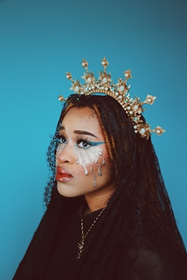 A person is adorned with a gold, ornate crown featuring intricate details and pearls. Their makeup includes striking silver tears and bold eye makeup, creating an artistic and dramatic appearance. A black lace veil partially covers their braided hair, and the background is a solid, calming blue.