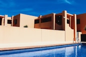 Modern building architecture with clean lines and geometric shapes, featuring warm beige walls contrasting against a clear blue sky. A pool in the foreground reflects the architecture and adds a sense of leisure and luxury.