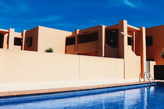 Modern building architecture with clean lines and geometric shapes, featuring warm beige walls contrasting against a clear blue sky. A pool in the foreground reflects the architecture and adds a sense of leisure and luxury.