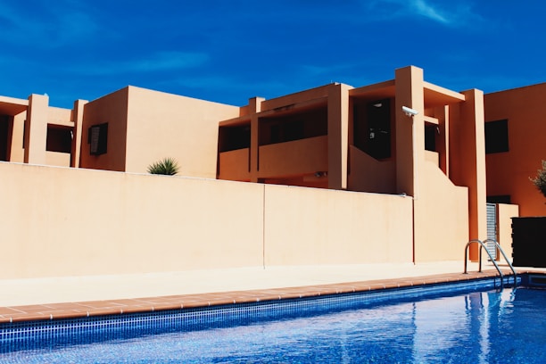 Modern building architecture with clean lines and geometric shapes, featuring warm beige walls contrasting against a clear blue sky. A pool in the foreground reflects the architecture and adds a sense of leisure and luxury.