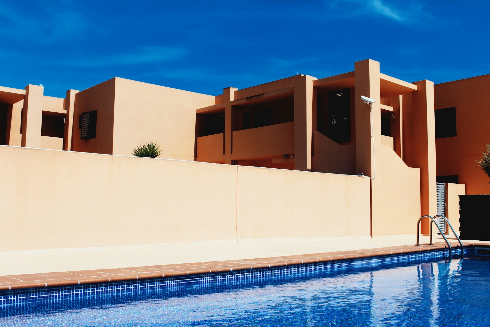 Modern building architecture with clean lines and geometric shapes, featuring warm beige walls contrasting against a clear blue sky. A pool in the foreground reflects the architecture and adds a sense of leisure and luxury.
