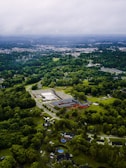Aerial view of a rural area with a mix of residential buildings, a school-like complex, and abundant green spaces. A dense forest surrounds the area, and several pools are visible near the houses. The horizon showcases a distant cityscape under a cloudy sky.