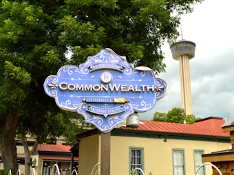 A decorative blue sign with ornate white details displays the text 'Commonwealth Coffeehouse & Bakery' along with the address. In the background, the top of a tall tower is visible against a cloudy sky, surrounded by lush green trees and a building with a red roof.