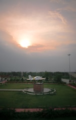A model airplane is mounted on a pedestal surrounded by a lush green park. The sun is setting or rising, casting a warm glow across a partly cloudy sky. There are pathways lined with greenery leading to the center display.