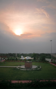 A model airplane is mounted on a pedestal surrounded by a lush green park. The sun is setting or rising, casting a warm glow across a partly cloudy sky. There are pathways lined with greenery leading to the center display.