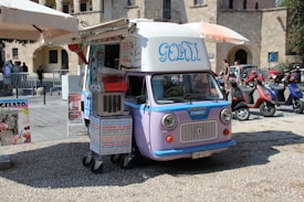 A vintage ice cream van with a cream and purple color scheme is parked on a cobblestone street. The van has a sign with the word 'Gelati' written in blue cursive letters on its side. It is equipped with an umbrella and signs advertising homemade ice cream in various languages. The background features several scooters parked along the street and people walking near a historical stone building.