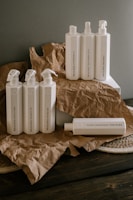 A collection of white plastic cleaning bottles with black text, arranged on crumpled brown paper and a dark wooden surface. The bottles have various labels such as 'Glass Mirror Steel Tincture' and 'Floor Concentrate Tincture.' The background is a plain gray wall.