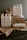 A collection of white plastic cleaning bottles with black text, arranged on crumpled brown paper and a dark wooden surface. The bottles have various labels such as 'Glass Mirror Steel Tincture' and 'Floor Concentrate Tincture.' The background is a plain gray wall.