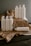 A collection of white plastic cleaning bottles with black text, arranged on crumpled brown paper and a dark wooden surface. The bottles have various labels such as 'Glass Mirror Steel Tincture' and 'Floor Concentrate Tincture.' The background is a plain gray wall.