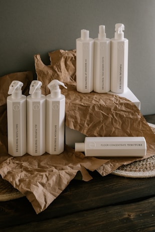 A collection of white plastic cleaning bottles with black text, arranged on crumpled brown paper and a dark wooden surface. The bottles have various labels such as 'Glass Mirror Steel Tincture' and 'Floor Concentrate Tincture.' The background is a plain gray wall.