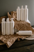 A collection of white plastic cleaning bottles with black text, arranged on crumpled brown paper and a dark wooden surface. The bottles have various labels such as 'Glass Mirror Steel Tincture' and 'Floor Concentrate Tincture.' The background is a plain gray wall.