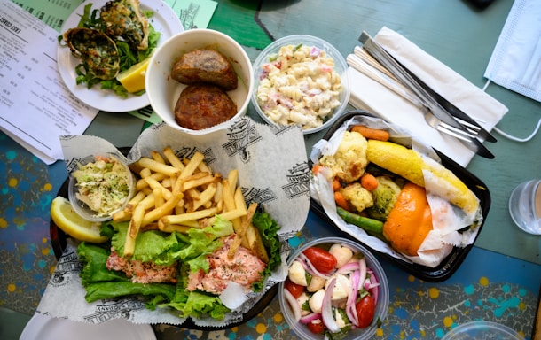 A table is set with a variety of dishes, including a plate of fries and a sandwich topped with lettuce and what seems to be a seafood filling. Surrounding it are sides of coleslaw and pasta salad, as well as a container with grilled vegetables like corn, carrots, and bell peppers. Nearby, there is a fresh salad with cherry tomatoes, mozzarella, and onions. Additional items include a plate of shellfish and meatballs, a menu, utensils wrapped in napkins, and a face mask placed on the table.