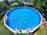 A large, round above-ground swimming pool fills most of the frame, its clear blue water reflecting sunlight. Several colorful pool noodles are floating at one end. The pool is set in a backyard with lush greenery and a trampoline is visible to one side. A wooden shed with solar panels on the roof is situated in the background.