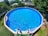 A large, round above-ground swimming pool fills most of the frame, its clear blue water reflecting sunlight. Several colorful pool noodles are floating at one end. The pool is set in a backyard with lush greenery and a trampoline is visible to one side. A wooden shed with solar panels on the roof is situated in the background.