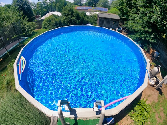 A large, round above-ground swimming pool fills most of the frame, its clear blue water reflecting sunlight. Several colorful pool noodles are floating at one end. The pool is set in a backyard with lush greenery and a trampoline is visible to one side. A wooden shed with solar panels on the roof is situated in the background.