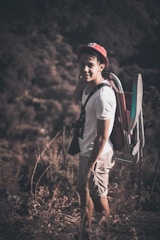 A person wearing a hat stands outdoors in a natural setting, smiling and carrying a foldable chair on their back. The individual is dressed casually in a white t-shirt and khaki shorts, with a pair of binoculars around their neck. The background consists of blurred greenery and shrubbery, suggesting a hiking or nature exploration scene.