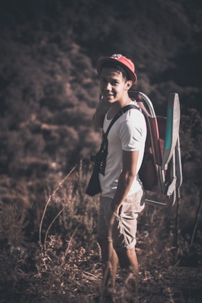 A person wearing a hat stands outdoors in a natural setting, smiling and carrying a foldable chair on their back. The individual is dressed casually in a white t-shirt and khaki shorts, with a pair of binoculars around their neck. The background consists of blurred greenery and shrubbery, suggesting a hiking or nature exploration scene.
