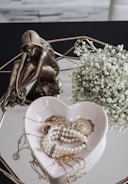 A bronze statuette of a seated, contemplative woman is positioned next to a delicate arrangement of white baby's breath flowers. In front of them, a heart-shaped ceramic dish holds a collection of jewelry, including pearl bracelets and golden accessories, against a reflective surface.