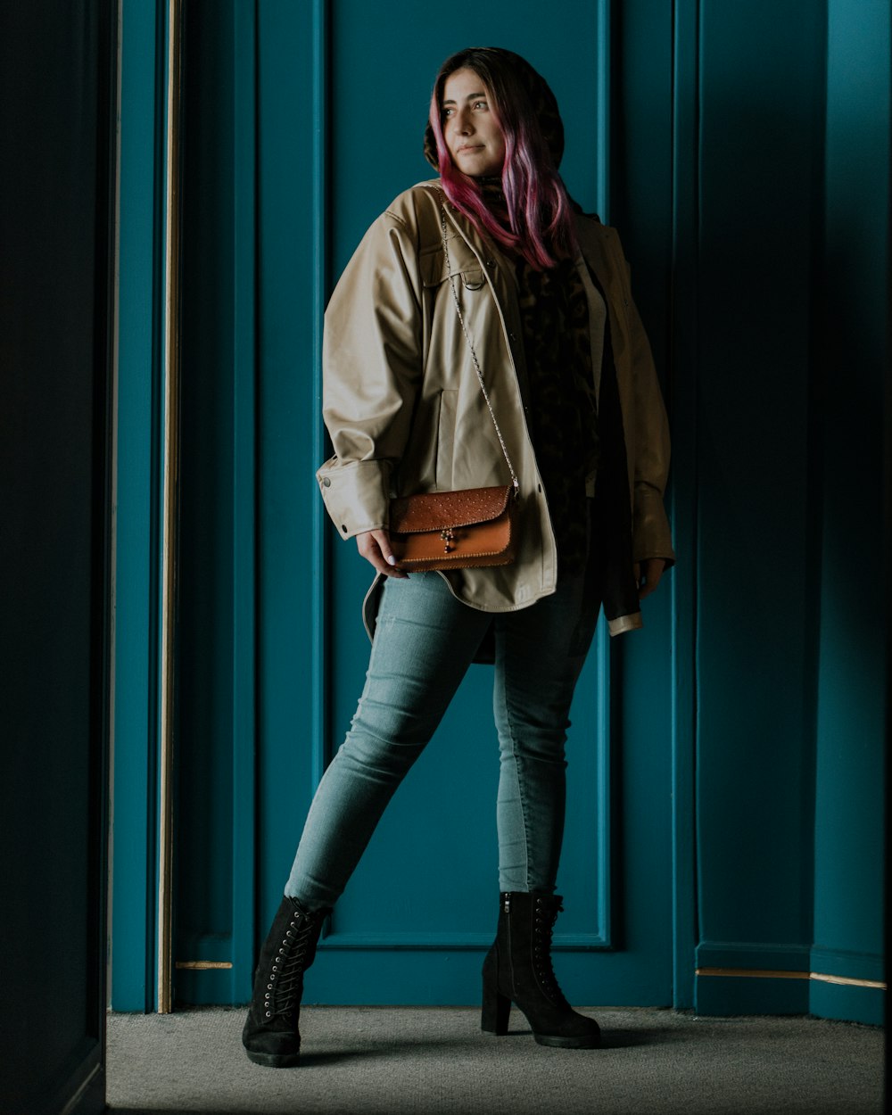 Mujer con abrigo marrón y jeans de mezclilla azul de pie junto a la pared verde y azul