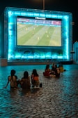 A large outdoor screen displays a soccer match at night. A small group of people sit on a cobblestone surface, watching the game. The screen is surrounded by a bright blue glow, creating a lively atmosphere.