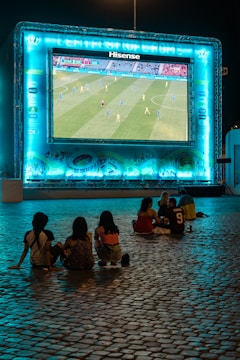 A large outdoor screen displays a soccer match at night. A small group of people sit on a cobblestone surface, watching the game. The screen is surrounded by a bright blue glow, creating a lively atmosphere.