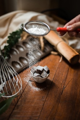 A hand holds a fine mesh sieve, sprinkling powdered sugar over a small bowl of chocolate truffles on a wooden table. Various baking utensils like a whisk, a rolling pin, and a baking tray are arranged around the scene. There's also a blurred background fabric draped casually.