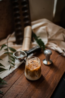 A glass of iced coffee with milk is placed on a rustic wooden surface. Nearby, there is a metal coffee tamper and an ice cream scooper. The background features a soft, white cloth draped over a surface, with dark green leaves scattered around, creating a cozy and inviting atmosphere.
